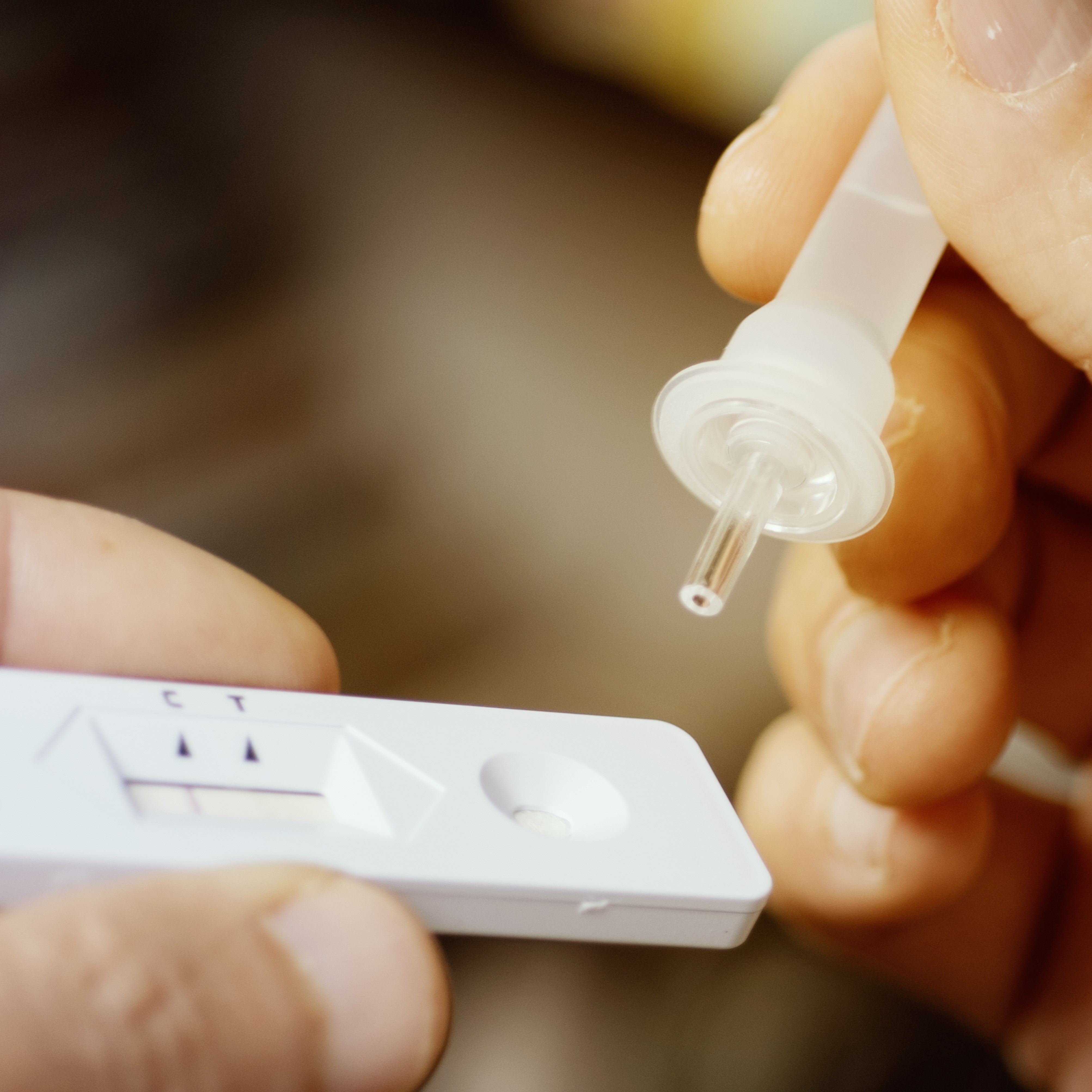 A person holds a thin, clear dropper above a test device. The test device features a rectangular shape with two indication lines. The setup suggests a process related to testing, likely for health-related purposes.