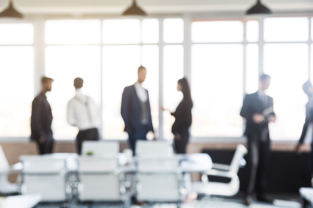 Eine Gruppe von fünf Personen in formeller Kleidung steht in einem hellen, modernen Büro und unterhält sich. Im Vordergrund sind Büromöbel sichtbar, während große Fenster Tageslicht hereinlassen. Die Szene vermittelt eine geschäftliche Atmosphäre.
