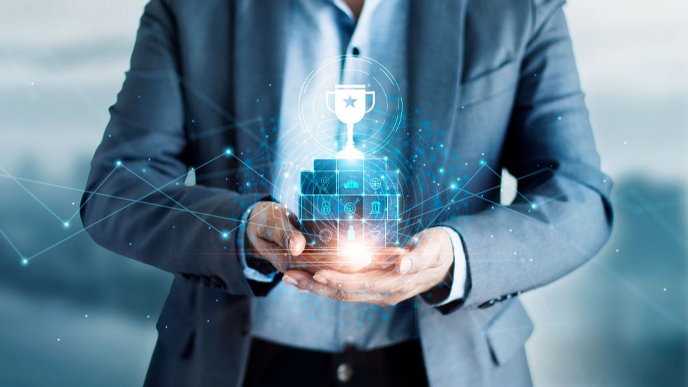 A person in a suit holds a glowing, digital trophy above a stack of icons representing achievement and data. The image conveys themes of success, innovation, and technology. Bright light and abstract lines enhance the futuristic feel.