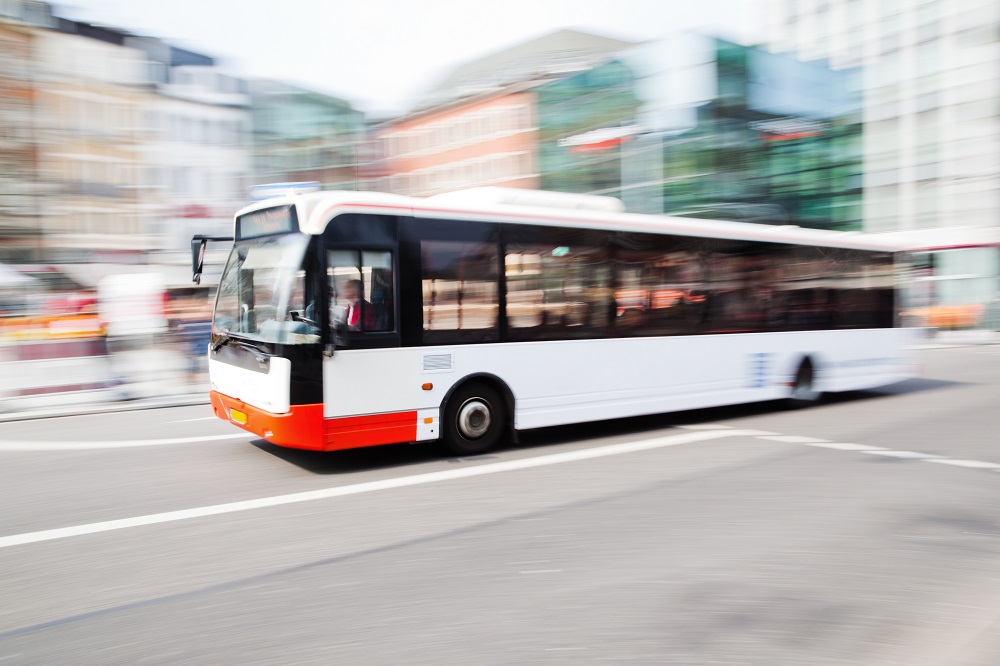A motion-blurred white and orange city bus travels along a street, suggesting speed. The background features buildings typical of an urban environment, with no discernible details due to the blur, emphasizing the bus as the central focus of the image.
