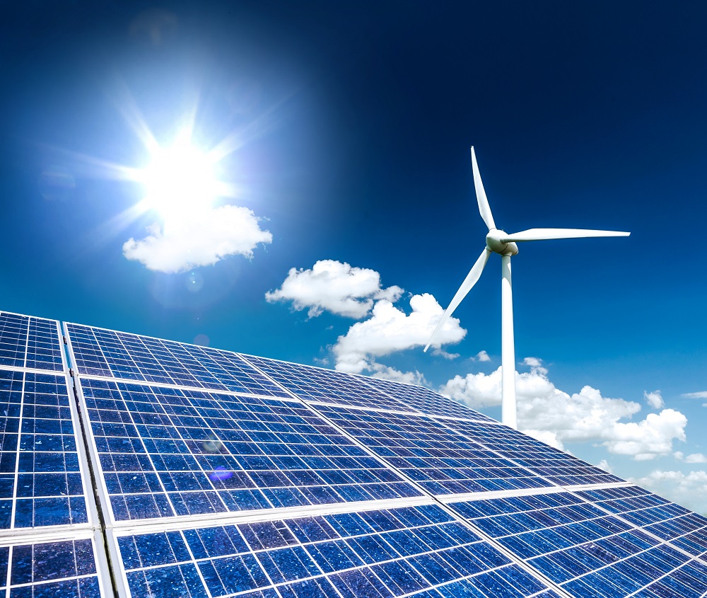 Ein Sonnenstrahl scheint auf Solarpanels, während im Hintergrund ein Windrad dreht. Der Himmel ist blau mit einigen weißen Wolken, was eine saubere und erneuerbare Energiequelle symbolisiert.