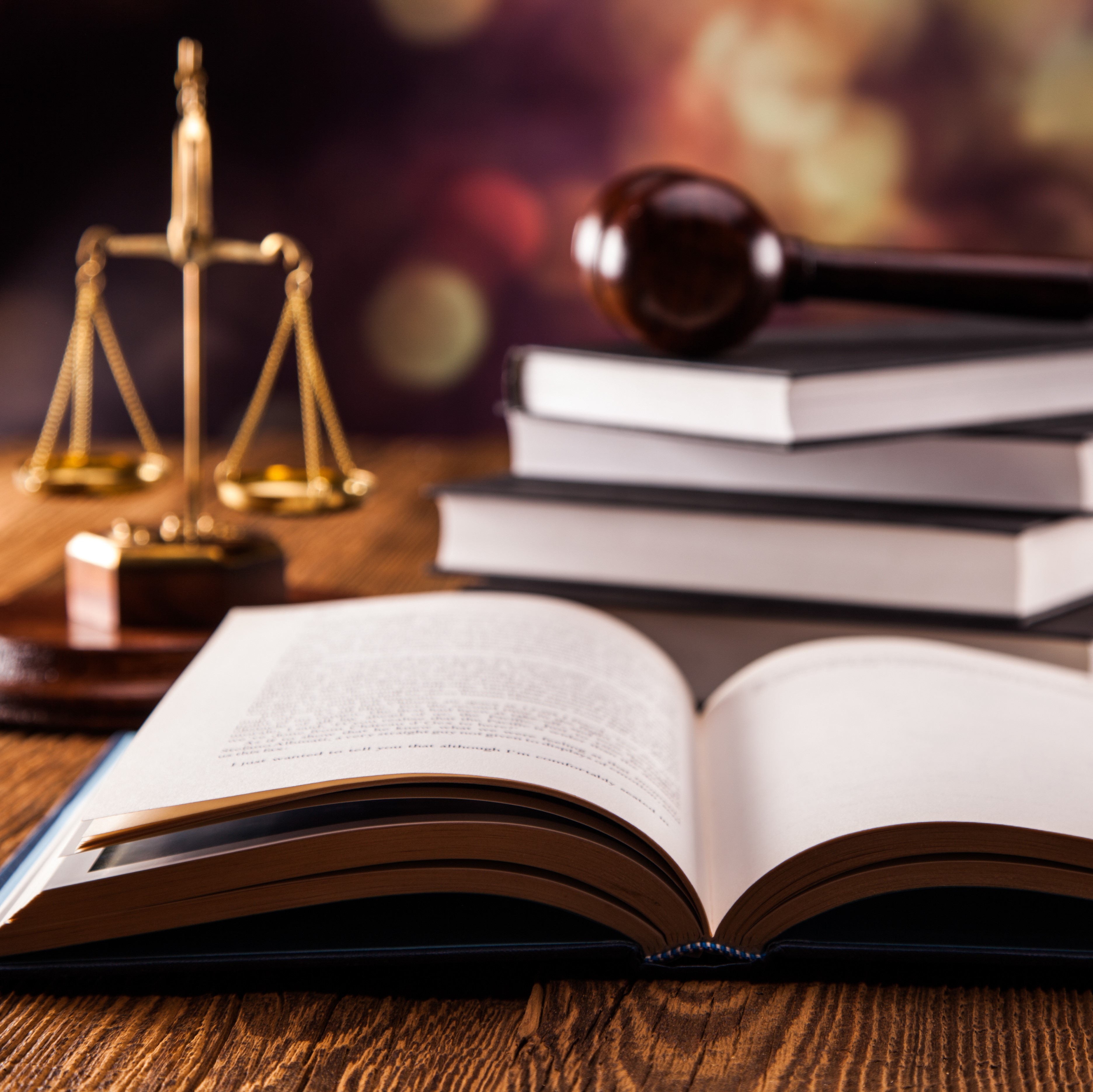 An open book rests on a wooden table, with its pages visible. Nearby, a stack of closed books is positioned alongside a brass scale symbolizing justice and a wooden gavel, representing legal themes and the judicial system.