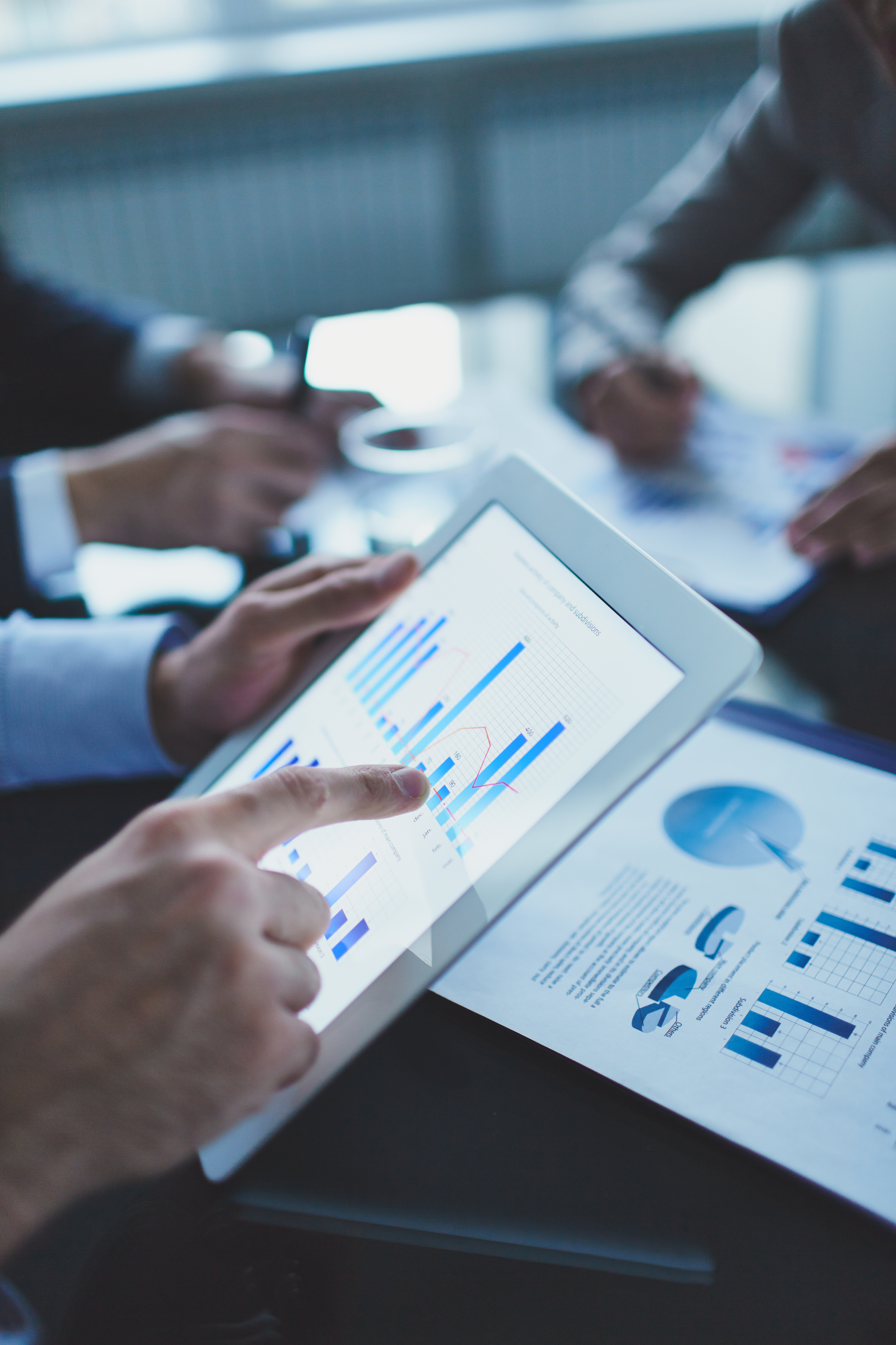 Eine Person zeigt mit dem Finger auf ein Tablet, das Diagramme und Datenvisualisierungen anzeigt. Im Hintergrund sind weitere Hände sichtbar, die Notizen machen und mit Dokumenten arbeiten. Die Szene suggeriert eine geschäftliche Besprechung zum Thema Datenanalyse.