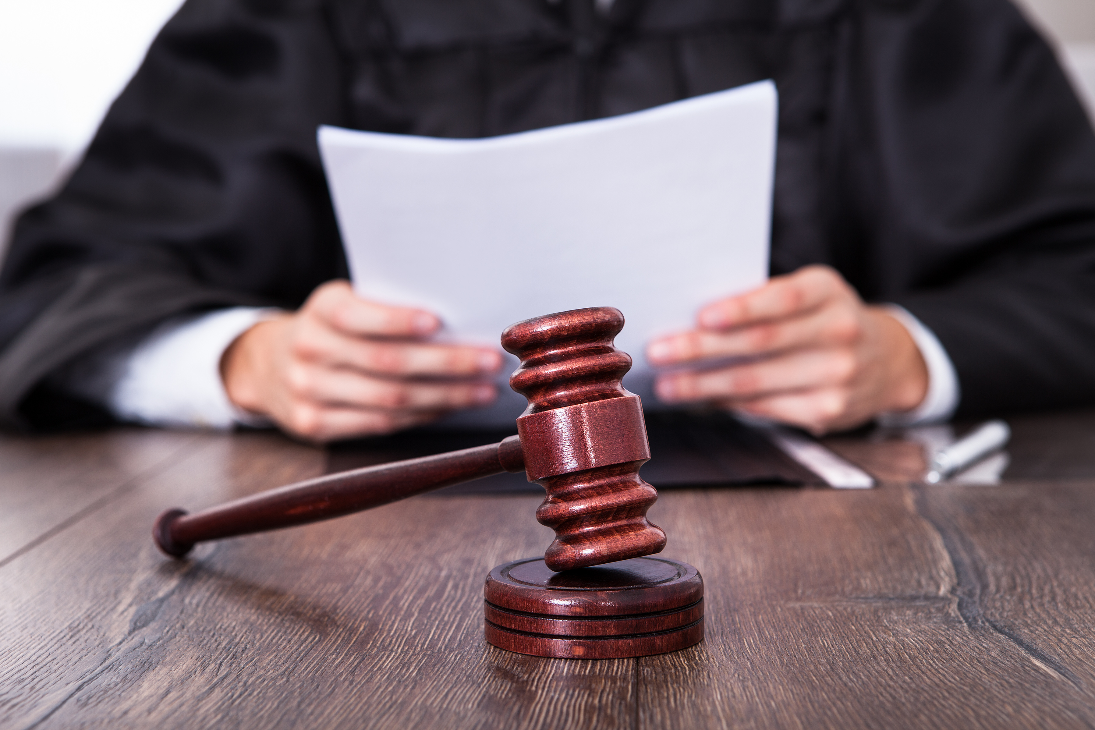 A judge in a black robe sits at a desk, holding a document, with a wooden gavel prominently displayed in the foreground. The setting suggests a courtroom environment, emphasizing judicial proceedings and decision-making.