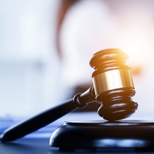 Ein Richterhammer liegt auf einem Tisch. Der Hammer hat einen goldenen Ring und symbolisiert Rechtsprechung und Justiz. Im Hintergrund ist ein verschwommener Bereich, der auf ein Gericht oder eine Rechtsumgebung hindeutet.