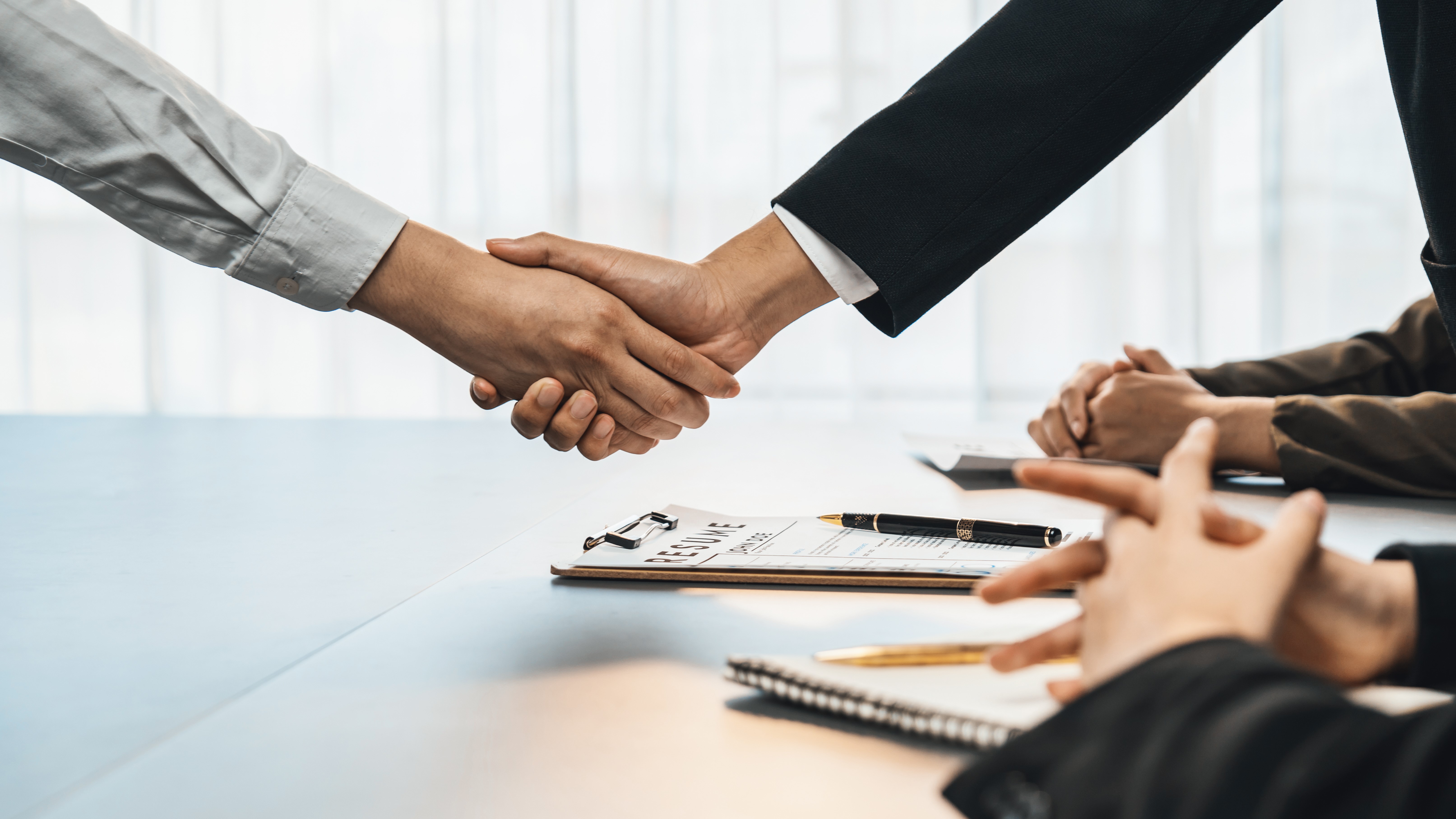 Zwei Personen geben sich in einem Geschäftstreffen die Hand. Im Vordergrund sind Hände von weiteren Teilnehmern zu sehen, die vor einem Tisch mit Notizen und Stiften sitzen. Das Bild vermittelt einen Moment der Einigung oder Zusammenarbeit.