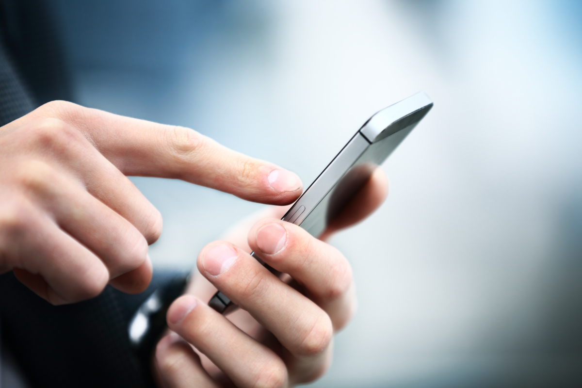 A close-up image of a hand holding a smartphone. The index finger is touching the screen, suggesting interaction. The background is blurred, emphasizing the hand and device.
