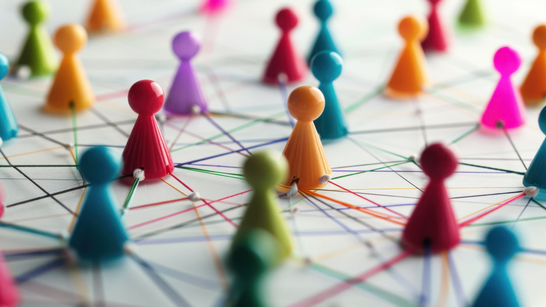 Eine Ansammlung von bunten Spielfiguren ist auf einer flachen Fläche angeordnet. Sie sind durch farbige Fäden miteinander verbunden, die ein Netzwerk bilden. Die Figuren symbolisieren Interaktionen oder Verbindungen zwischen Gruppen oder Individuen.