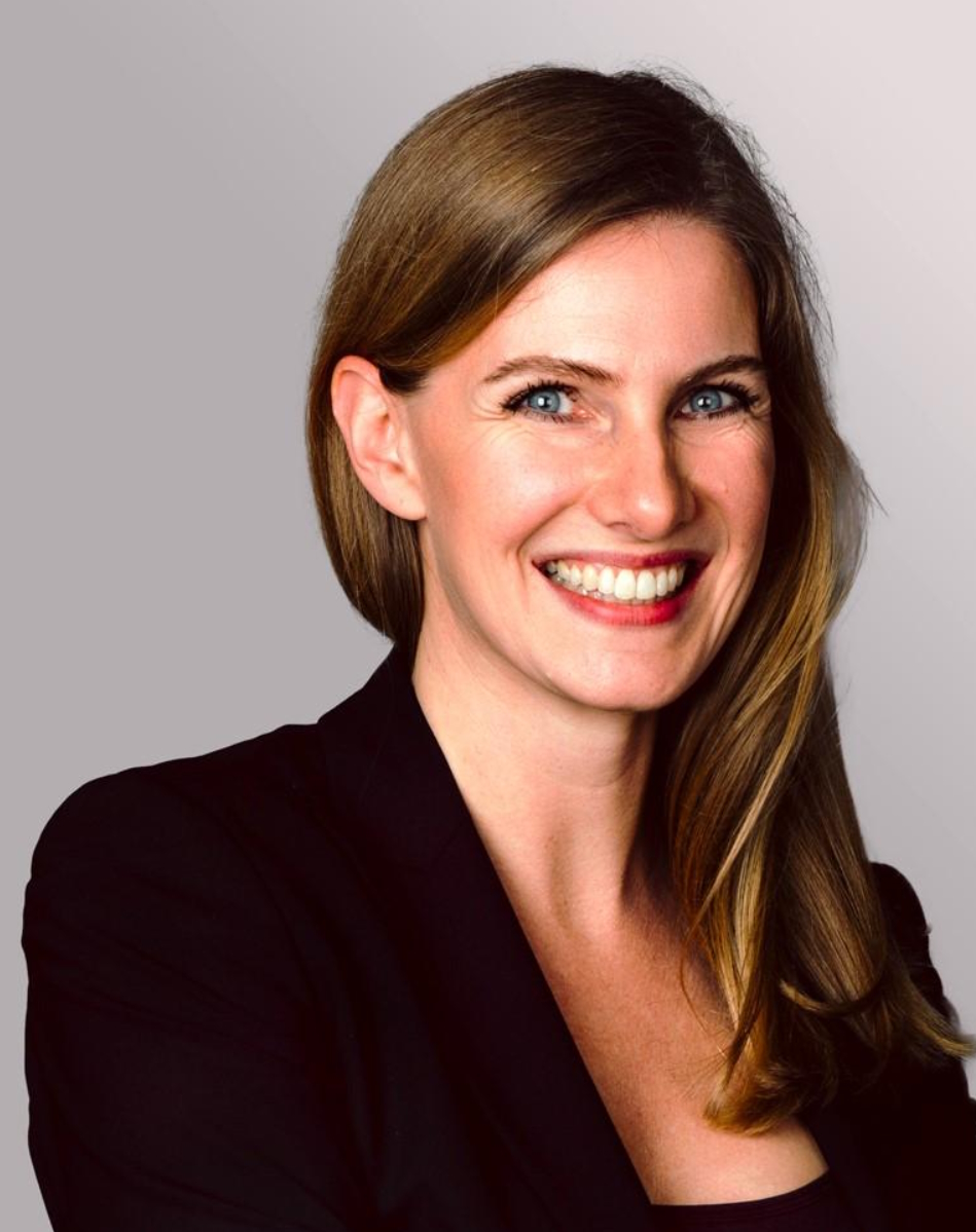 Porträt einer lächelnden Frau mit langen, blonden Haaren, die einen schwarzen Blazer trägt. Der Hintergrund ist in sanften Grautönen gehalten. Ihr Gesicht zeigt eine freundliche und offene Ausstrahlung.