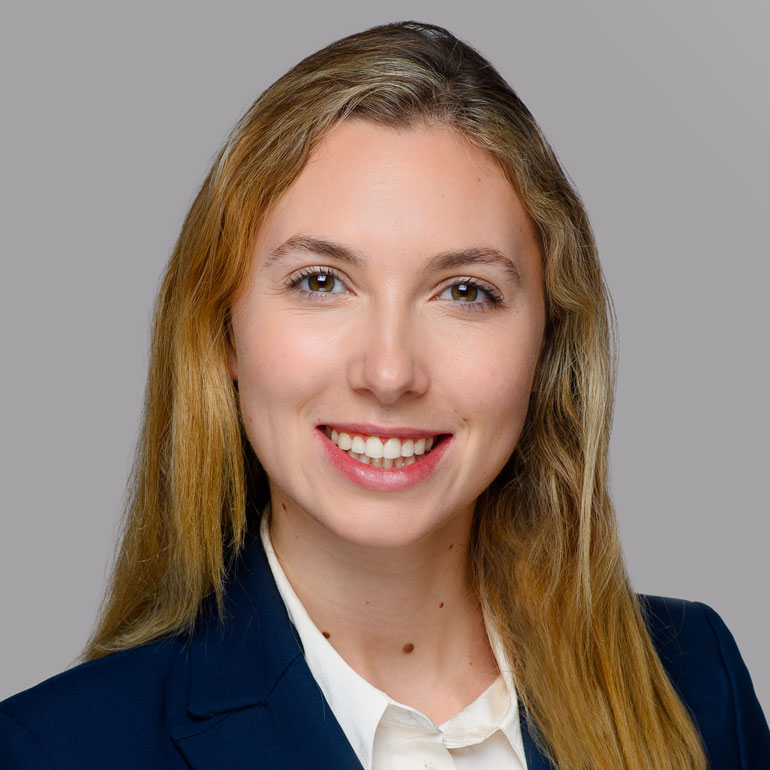 Porträt einer lächelnden Frau mit langen, blonden Haaren. Sie trägt einen dunklen Anzug und eine helle Bluse. Der Hintergrund ist grau. Die Frau wirkt freundlich und professionell.