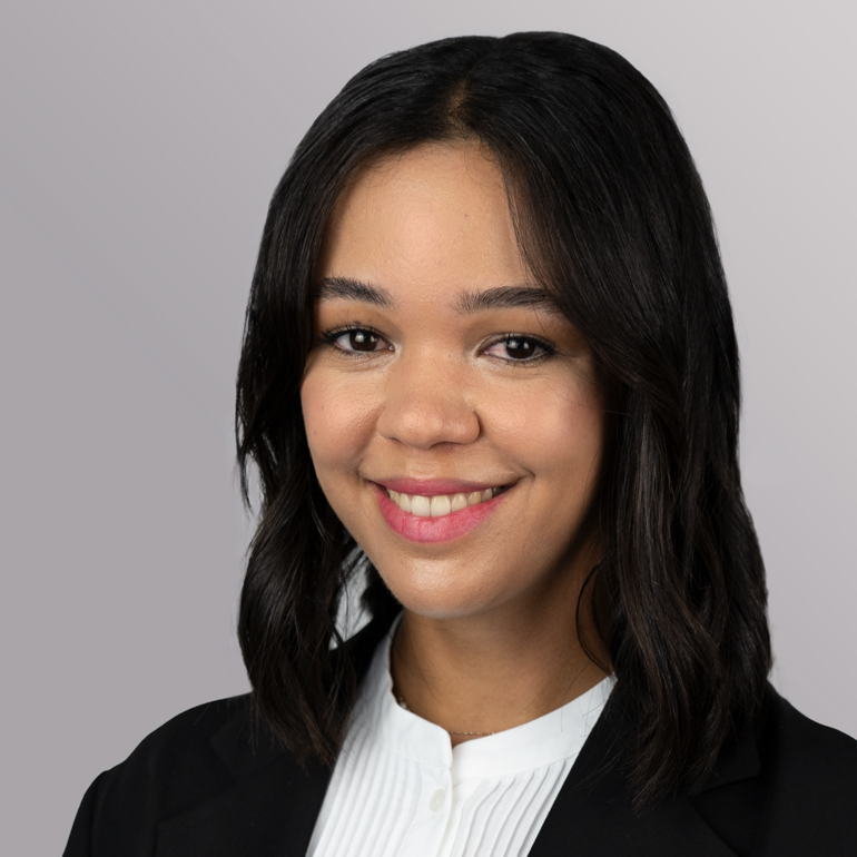 Eine junge Frau mit mittellangen, dunklen Haaren lächelt freundlich in die Kamera. Sie trägt ein weißes Hemd und einen schwarzen Blazer. Der Hintergrund ist sanft grau.