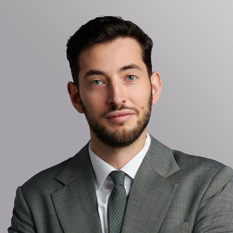 Ein Mann mit dunklen Haaren und einem kurzen Bart trägt einen grauen Anzug und eine grüne Krawatte. Er schaut direkt in die Kamera und hat einen neutralen Gesichtsausdruck. Der Hintergrund ist einfarbig in einem sanften Grauton.