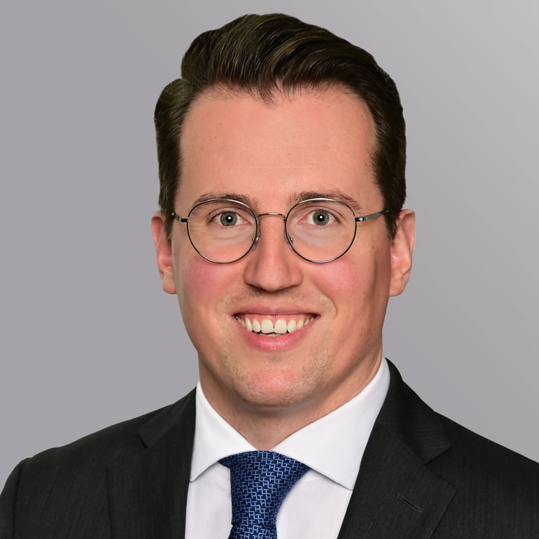 Porträt eines lächelnden Mannes mit einem Anzug, Krawatte und runden Brillen. Er hat kurzes, dunkles Haar und einen freundlichen Gesichtsausdruck. Der Hintergrund ist einfarbig grau.