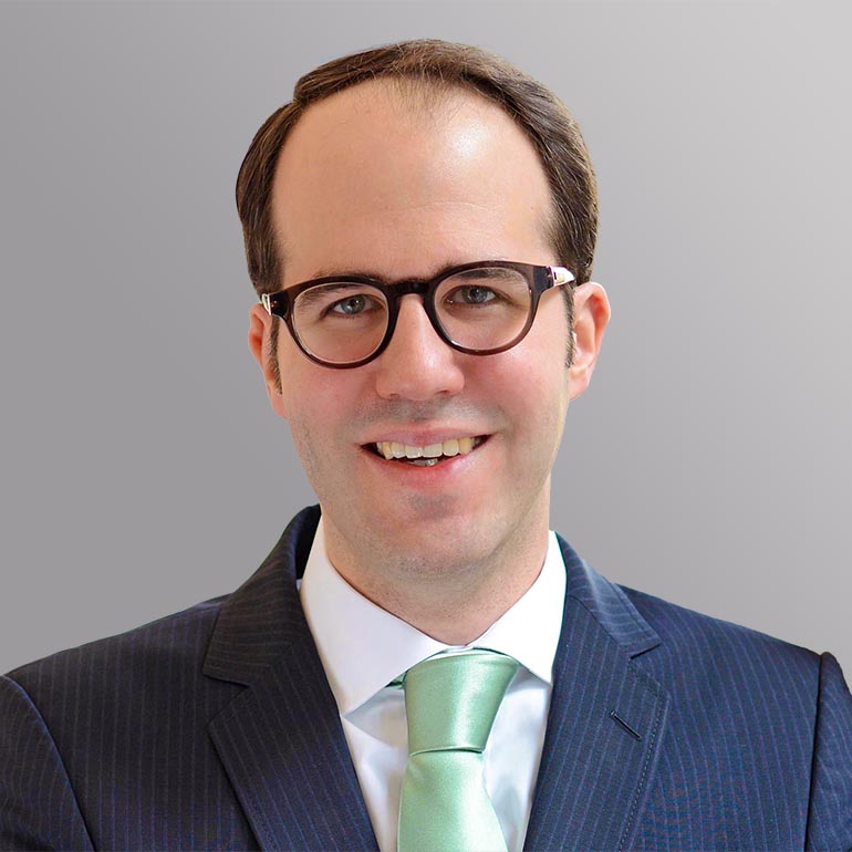 Porträt eines Mannes mit Brille, lächelnd. Er trägt einen dunkelblauen Anzug mit stripes und ein helles Hemd mit einer grünen Krawatte. Der Hintergrund ist grau.