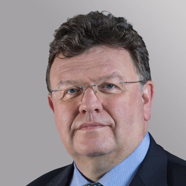 Ein mittelalter Mann mit kurzen, dunklen Haaren und Brille schaut direkt in die Kamera. Er trägt einen Anzug und ein hellblaues Hemd. Der Hintergrund ist grau. Sein Gesichtsausdruck wirkt neutral und selbstbewusst.