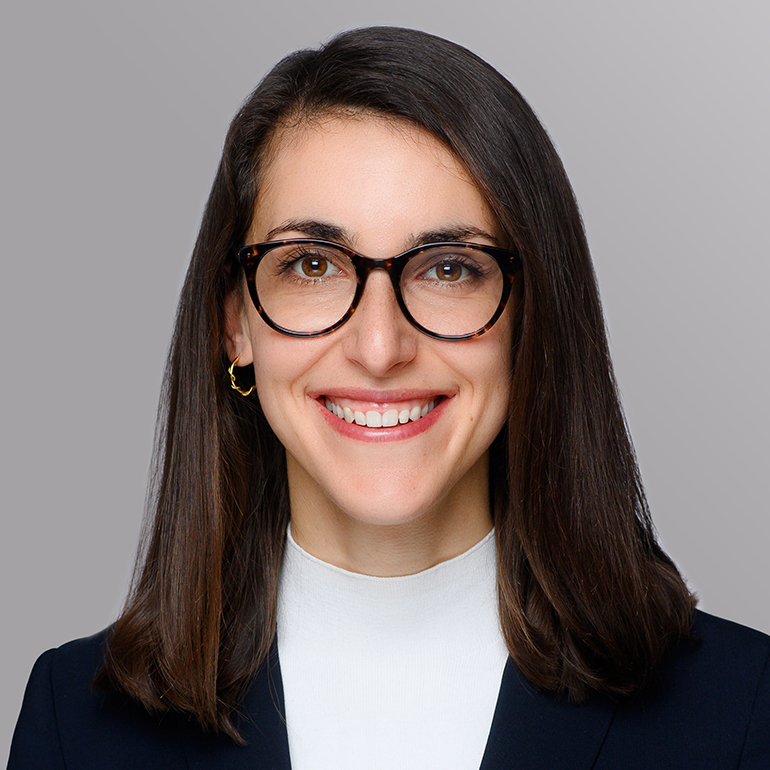 Ein Porträt einer lächelnden Frau mit langen, dunklen Haaren. Sie trägt eine weiße Bluse und einen dunkelblauen Blazer sowie runde, schwarze Brillen. Der Hintergrund ist grau und neutral gehalten, um den Fokus auf ihr Gesicht zu lenken.