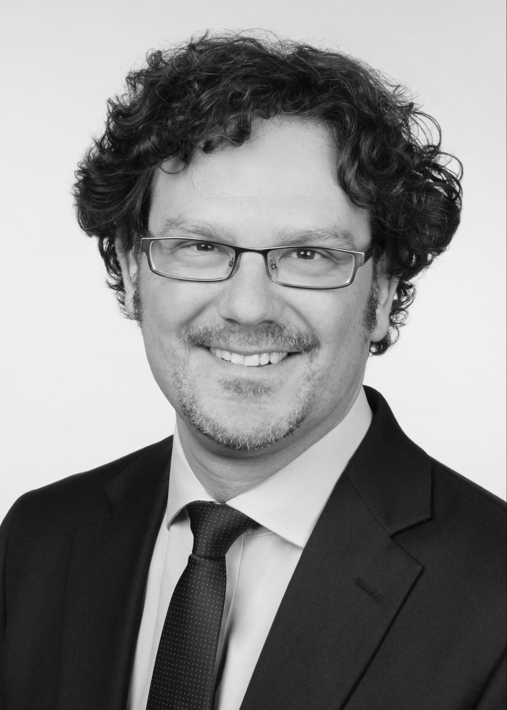 Porträt eines Mannes mit lockigem, dunklem Haar und Brille. Er trägt einen schwarzen Anzug und eine helle Krawatte mit Punkten. Er lächelt freundlich in die Kamera, vor einem weißen Hintergrund.