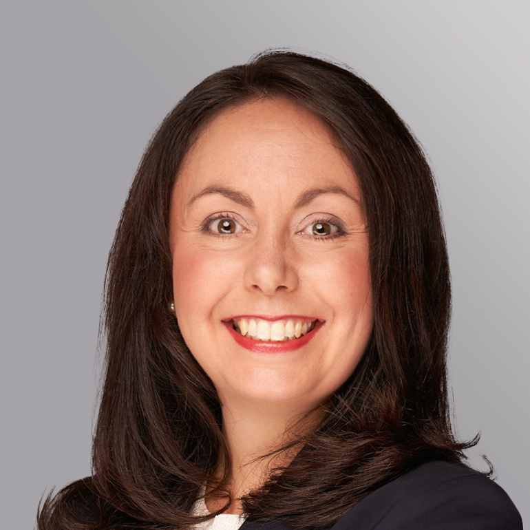 Porträt einer lächelnden Frau mit langen, dunklen Haaren und einem blauen Kleidungsstück. Der Hintergrund ist grau. Die Frau hat einen freundlichen Ausdruck und wirkt selbstbewusst.