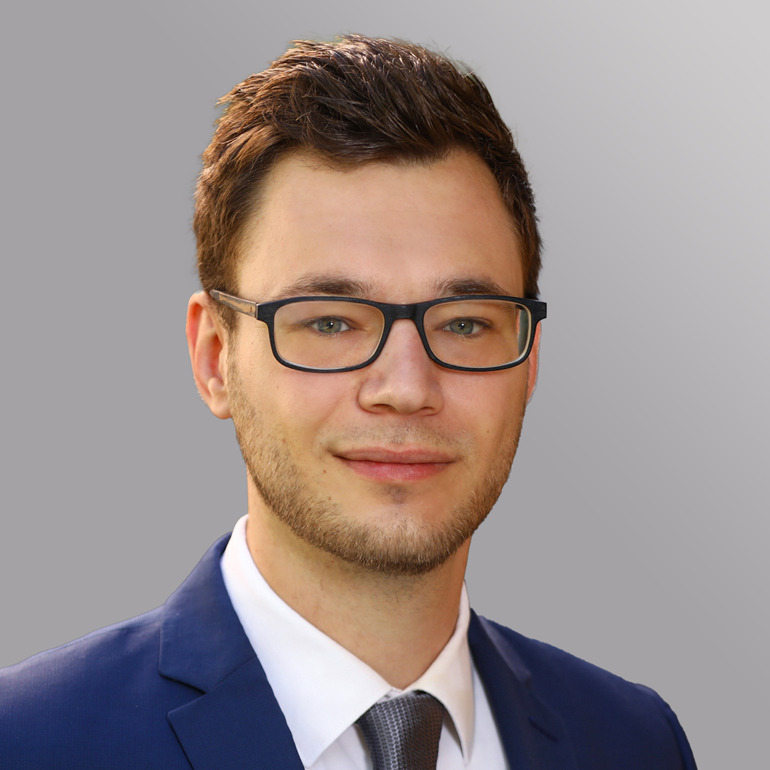 Ein junger Mann mit kurzen, dunklen Haaren und einem leichten Bart lächelt in die Kamera. Er trägt eine blaue Anzugjacke, ein weißes Hemd und eine graue Krawatte. Der Hintergrund ist einfarbig grau, was den Fokus auf sein Gesicht lenkt.