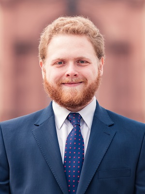 Ein junger Mann mit einem vollen Bart lächelt freundlich in die Kamera. Er trägt einen dunklen Anzug mit einer weiß-blauen Krawatte, die kleine rote Punkte hat. Der Hintergrund ist unscharf, zeigt jedoch eine architektonische Struktur.