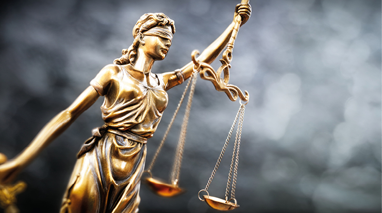 Eine goldene Statue der Justitia, weiblich, mit verbundenen Augen, die eine Waage in der rechten Hand hält. Die Waage symbolisiert Gerechtigkeit und Ausgewogenheit. Der Hintergrund ist verschwommen und unauffällig, um den Fokus auf die Figur zu lenken.