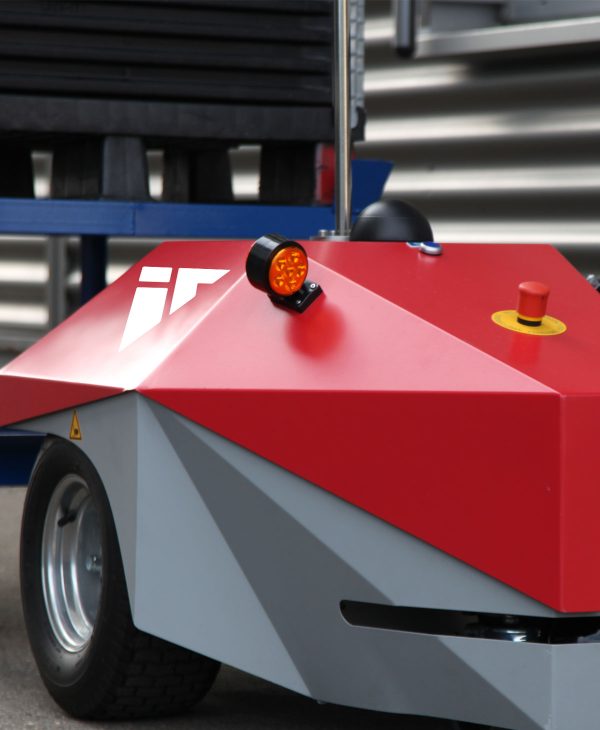 Ein roter, geometrisch geformter Roboter mit grauen Elementen ist abgebildet. Er hat ein gelbes Knopf und ein orangefarbenes Licht auf der Oberseite. Der Roboter steht auf zwei Reifen und hat eine moderne, futuristische Erscheinung.