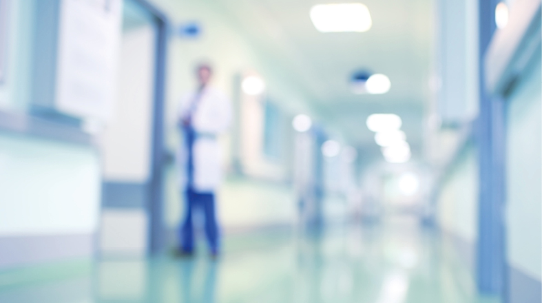 Das Bild zeigt einen unscharfen Krankenhausflur mit einem Arzt in einem weißen Kittel im Vordergrund. Die Wände sind hell und die Beleuchtung sorgt für eine sterile Atmosphäre. Der Flur wirkt lang und führt zu weiteren Türen, die nicht klar erkennbar sind.