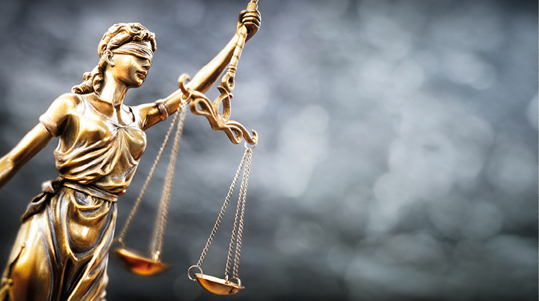 Eine goldene Justitia-Statue hält eine Waage in der einen Hand und ein Schwert in der anderen. Sie trägt eine Augenbinde, was Gerechtigkeit ohne Vorurteile symbolisiert. Der Hintergrund ist verschwommen und vermittelt einen neutralen, ernsthaften Eindruck.