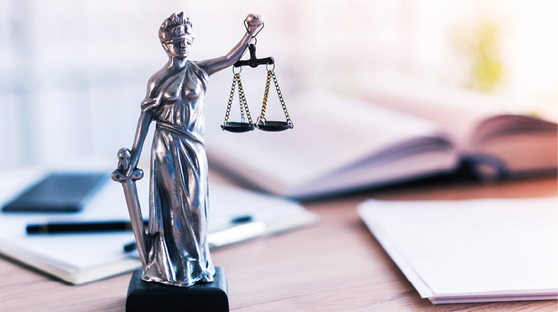 Eine silberne Statue von Justitia, der römischen Göttin der Gerechtigkeit, hält eine Waage in der rechten Hand und ein Schwert in der linken Hand. Sie steht auf einem Tisch, umgeben von Notizblock und Schreibutensilien, was ein rechtliches oder juristisches Umfeld symbolisiert.