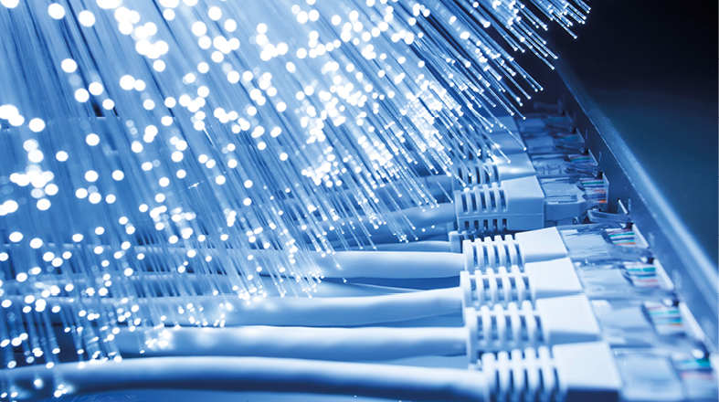 Das Bild zeigt eine Nahaufnahme von Glasfaserkabeln, die in einem Netzwerkteil angeschlossen sind. Die Fasern strahlen ein bläuliches Licht aus, während die Kabelstecker zusammengeordnete weiße Kabel darstellen, die zur Datenübertragung verwendet werden.