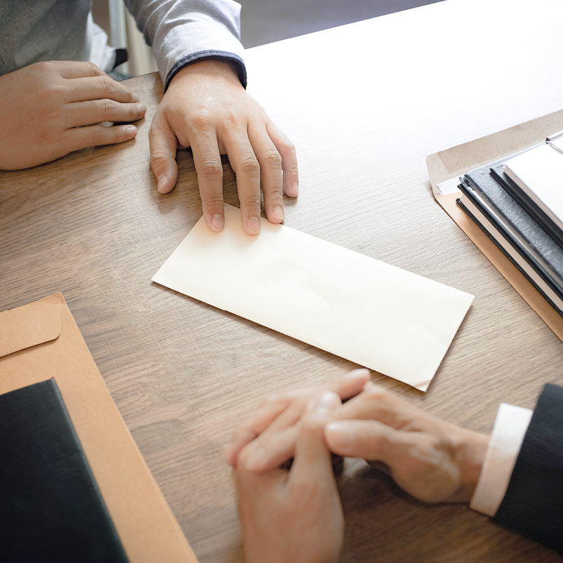 Zwei Personen sitzen an einem Tisch. Eine Hand liegt auf einem weißen Umschlag in der Mitte, während die andere Hand sanft die Hand des Gegenübers umfasst. Der Tisch ist mit verschiedenen Unterlagen und Aktenordnern dekoriert.
