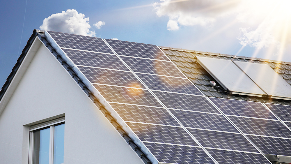 Das Bild zeigt die Dachfläche eines Hauses, die mit zahlreichen Solarpanels ausgestattet ist. Im Hintergrund sind ein blauer Himmel und einige Wolken zu sehen. Die Sonnenstrahlen reflektieren auf den Panels, was auf eine sonnige Umgebung hinweist.