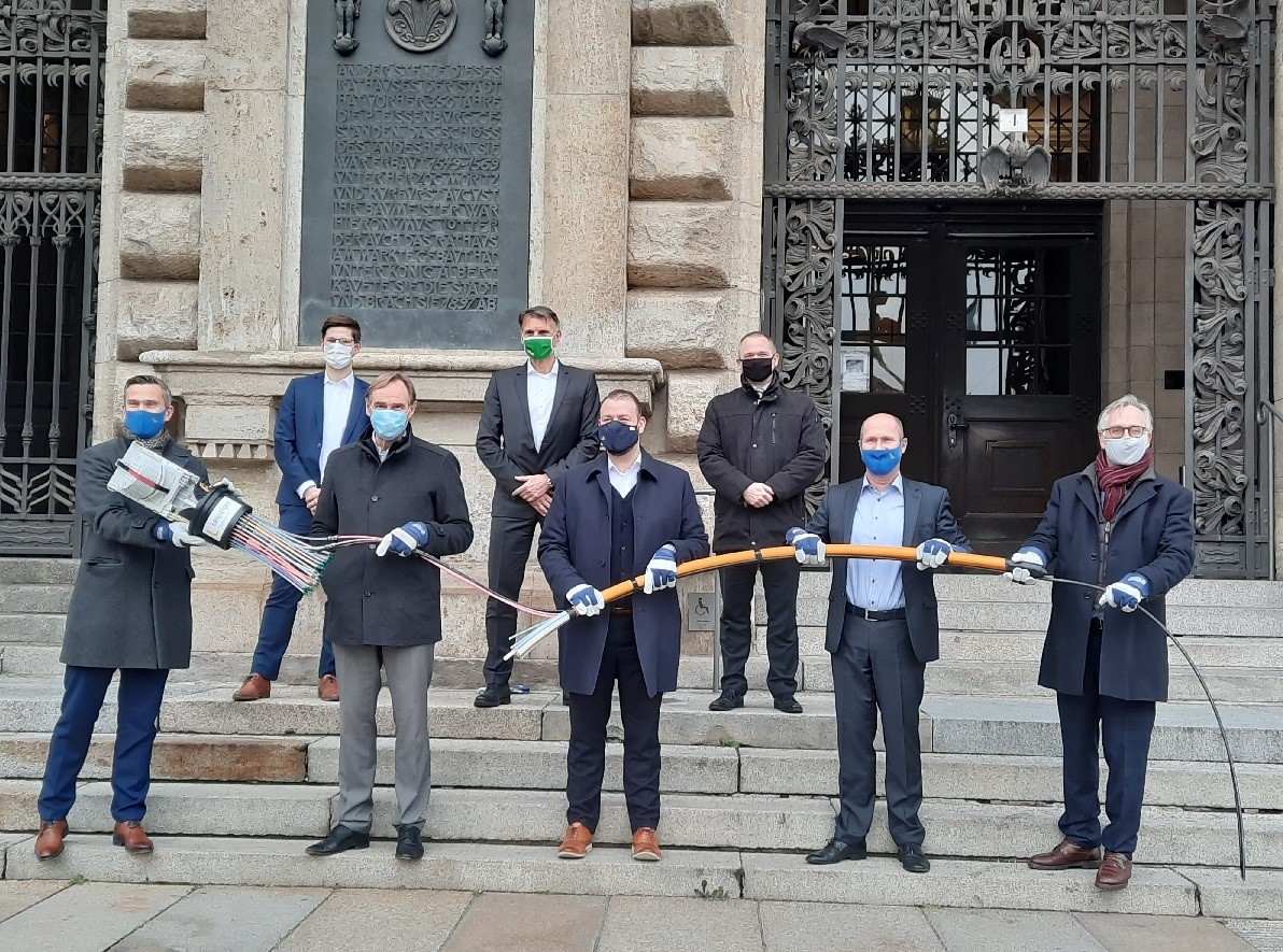 A group of seven individuals is gathered on the steps of a building, wearing masks. They hold various cables and equipment, suggesting a technology or connectivity initiative. The setting has historical architecture, emphasizing a formal occasion related to telecommunications.