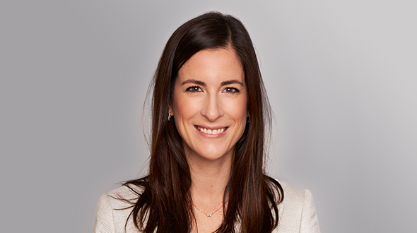 Ein Porträt einer lächelnden Frau mit langen, braunen Haaren. Sie trägt einen hellen Blazer und hat einer offenen Ausstrahlung. Der Hintergrund ist einfarbig und neutral, was die Aufmerksamkeit auf ihr Gesicht lenkt.