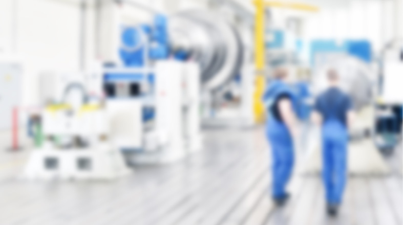 Zwei Personen in blauen Arbeitsanzügen gehen in einer Industriehalle, die mit Maschinen und technischen Geräten ausgestattet ist. Die Umgebung ist hell und modern, mit einem Fokus auf den technischen Arbeitsbereich.