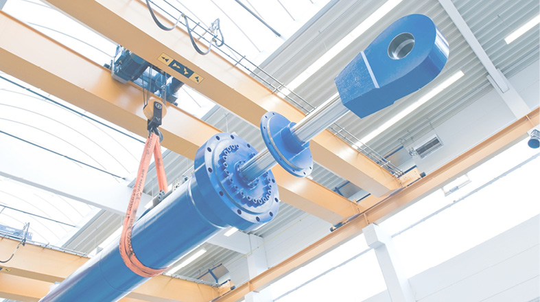 Das Bild zeigt eine technische Vorrichtung mit einem blauen Zylinder, der an einem Haken befestigt ist. Der Zylinder hat einen großen Verbindungsflansch und wird von einem Kran gehalten, der sich über ihm befindet. Die Umgebung sieht industriell aus, mit hohen Decken und hellem Licht.