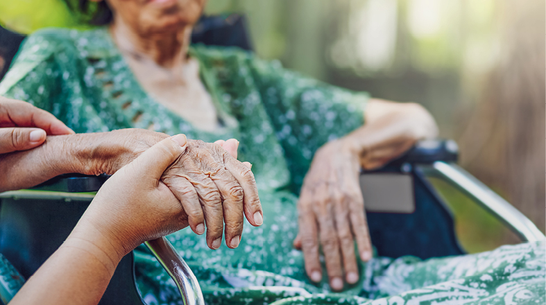 Eine ältere Frau sitzt im Rollstuhl und hält die Hand einer jüngeren Person. Die beiden Hände drücken Nähe und Zuneigung aus. Die ältere Dame trägt ein grünes Kleid, während der Hintergrund unscharf ist, was die emotionale Verbindung betont.