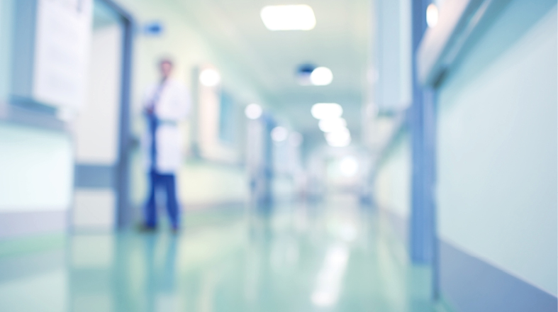 Ein verschwommenes Bild eines Krankenhausflurs mit einem Arzt im Hintergrund. Der Flur ist hell erleuchtet und zeigt medizinische Einrichtungen. Der Fokus liegt auf der Präsenz des Arztes, der anscheinend einer Patientensituation angehört.