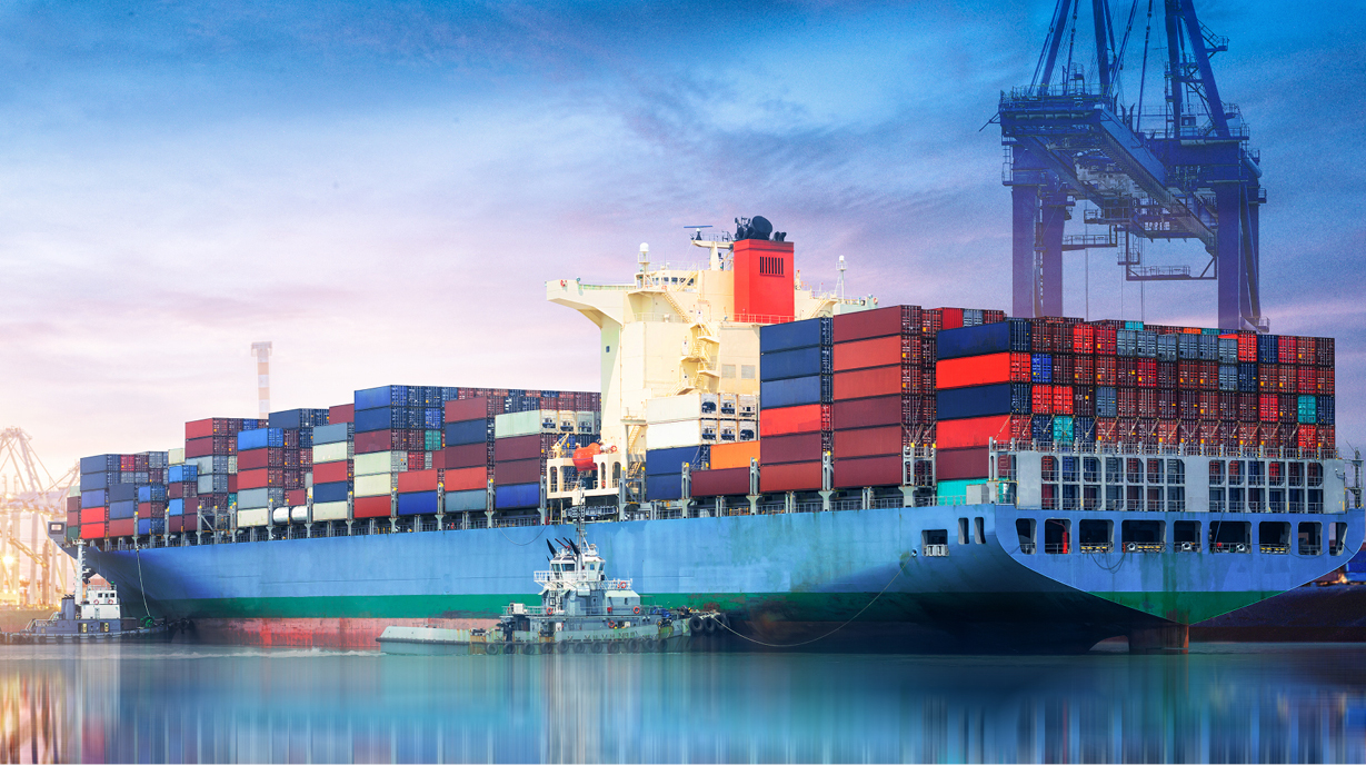 Ein großes Containerschiff liegt im Hafen, beladen mit zahlreichen bunten Container. Im Hintergrund sind Kräne und der Himmel ist in sanften Farben von Sonnenaufgang oder Sonnenuntergang getaucht.