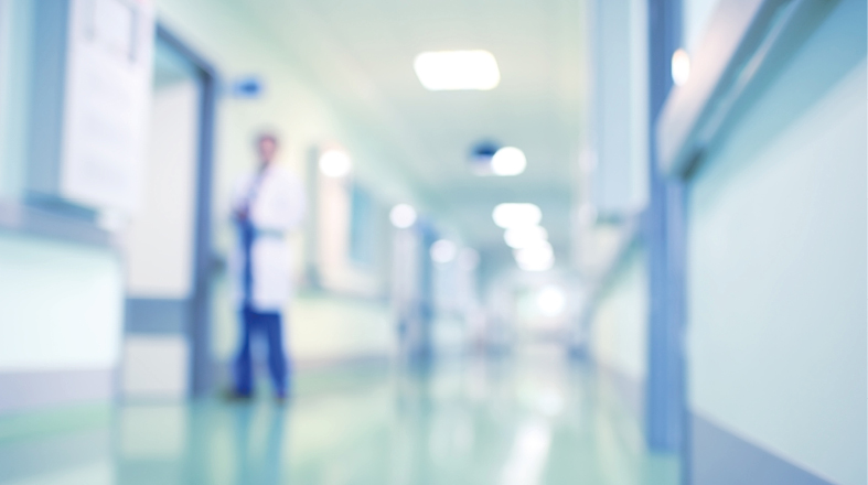 Ein verschwommener Krankenhausflur mit einem Arzt im Hintergrund. Der Flur ist hell beleuchtet und zeigt mehrere Türen zu Behandlungsräumen. Der Fokus liegt auf der Präsenz des Arztes und der medizinischen Umgebung.