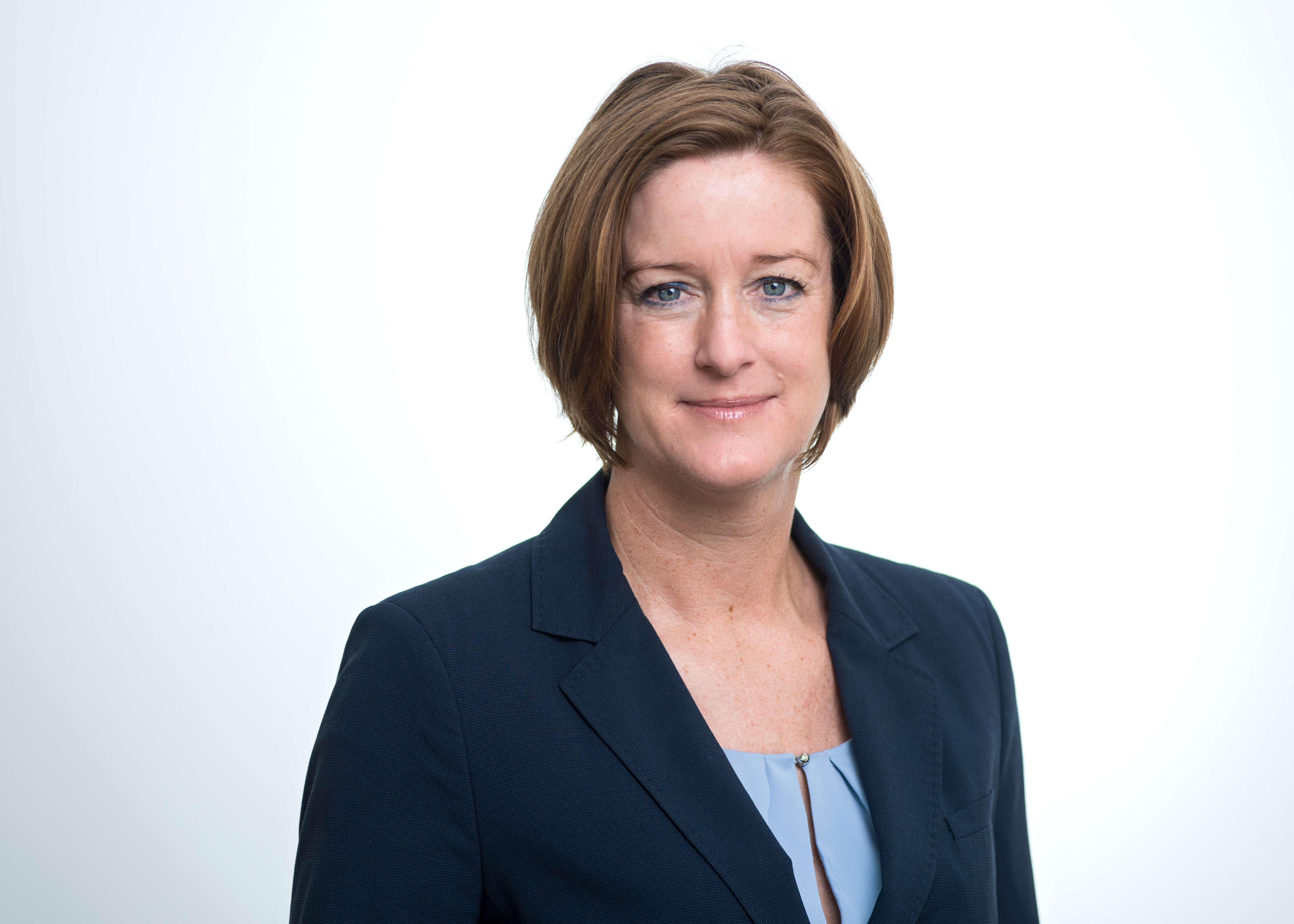A professional woman smiles confidently, wearing a dark blazer over a light blouse. Her short, brown hair frames her face, which is set against a bright, neutral background. The expression conveys approachability and professionalism.
