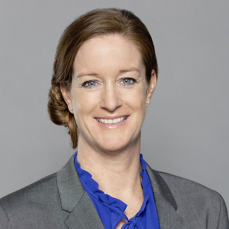 Porträt einer Frau mit lächelndem Gesicht. Sie hat braune Haare, die zu einem niedrigen Dutt frisiert sind, und trägt eine graue Blazerjacke sowie ein blaues Oberteil mit Rüschen. Der Hintergrund ist hellgrau, was den Fokus auf ihr Gesicht lenkt.