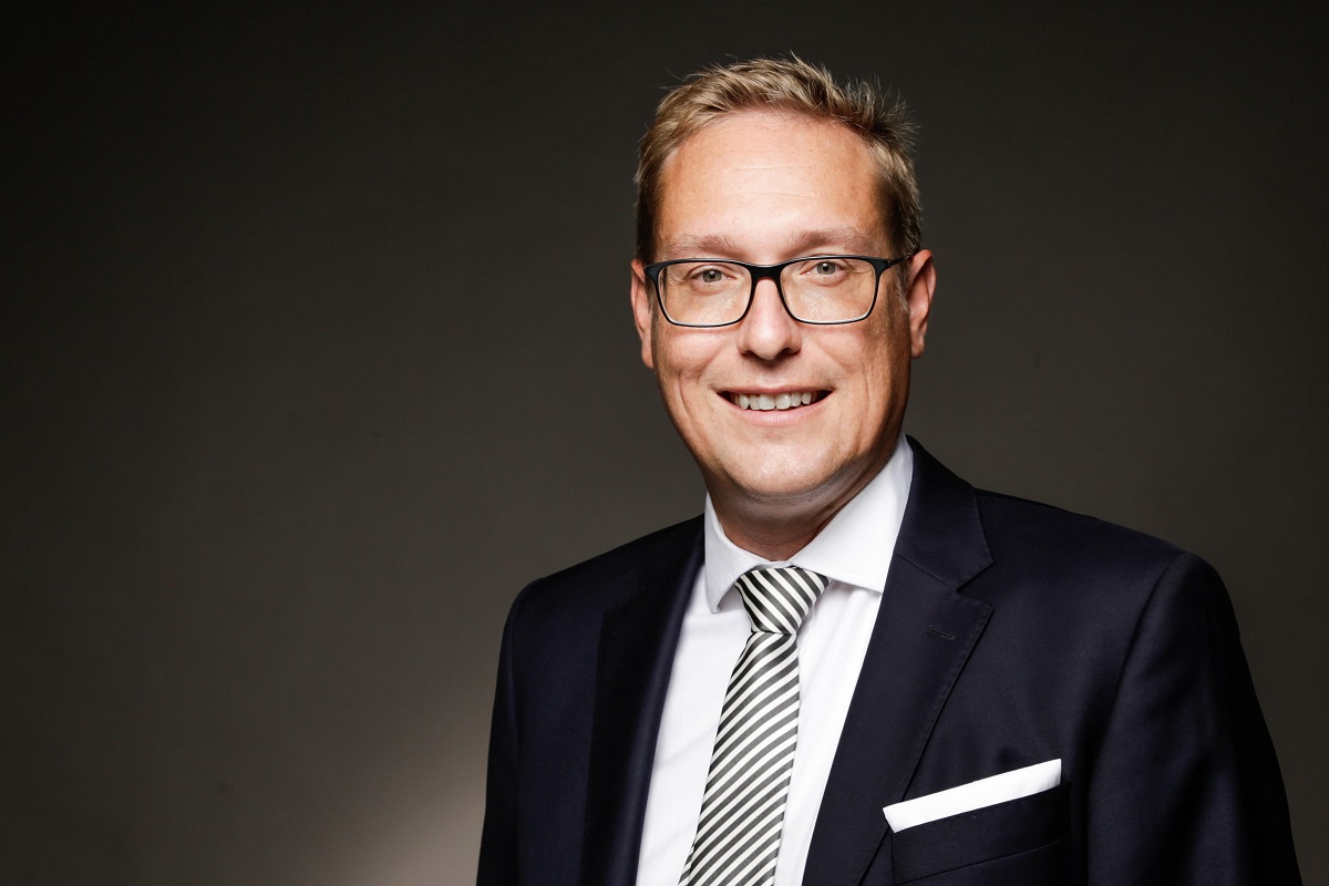 A middle-aged man smiles confidently at the camera, wearing glasses and a dark suit with a striped tie. He has short, light brown hair and stands against a dark background, projecting a professional and approachable demeanor.