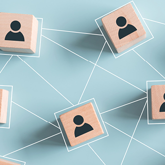 Die Grafik zeigt Holzblöcke mit stilisierten Personen-Symbolen, die durch Linien verbunden sind. Dies symbolisiert Netzwerkverbindungen oder die Interaktion zwischen Individuen in einem sozialen oder geschäftlichen Kontext. Der Hintergrund ist hellblau.