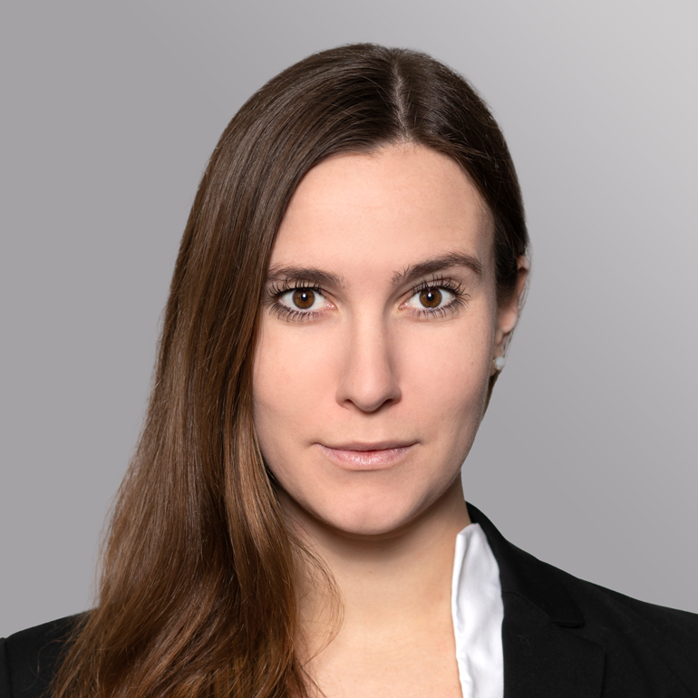 Das Bild zeigt eine Frau mit langen, braunen Haaren und einem neutralen Gesichtsausdruck. Sie trägt ein schwarzes Blazer-Oberteil und eine weiße Bluse. Der Hintergrund ist grau und weich, wodurch der Fokus auf ihrem Gesicht liegt.