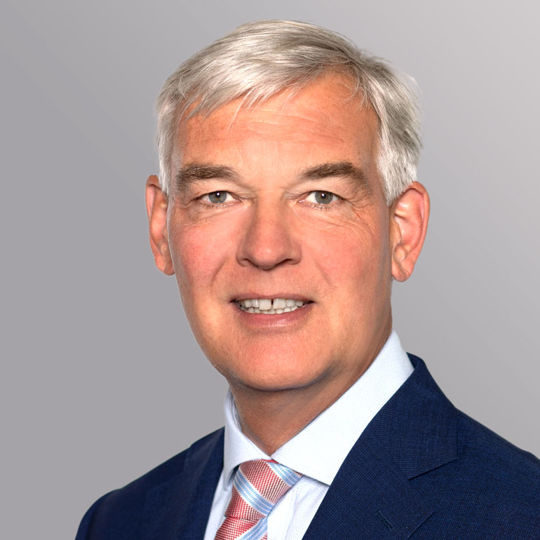 Porträt eines Mannes mit grauen Haaren und einem freundlichen Lächeln. Er trägt einen blauen Anzug und ein kariertes Hemd mit einer Krawatte. Der Hintergrund ist hellgrau, was das Gesicht hervorhebt.