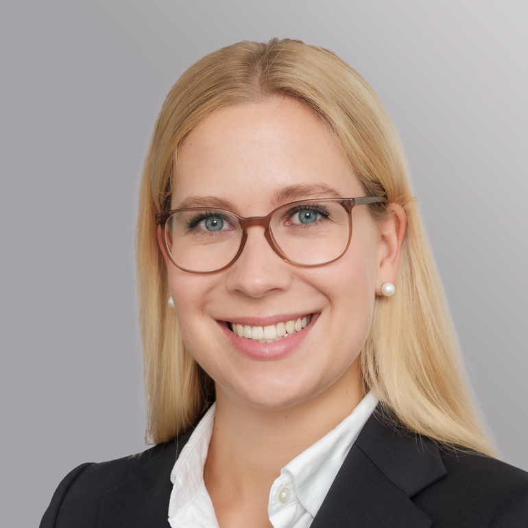 Das Bild zeigt eine lächelnde Frau mit langen blonden Haaren, die eine Brille trägt. Sie hat einen blauen Blazer und eine weiße Bluse an. Der Hintergrund ist grau und weich, was ihr freundliches Gesicht betont.