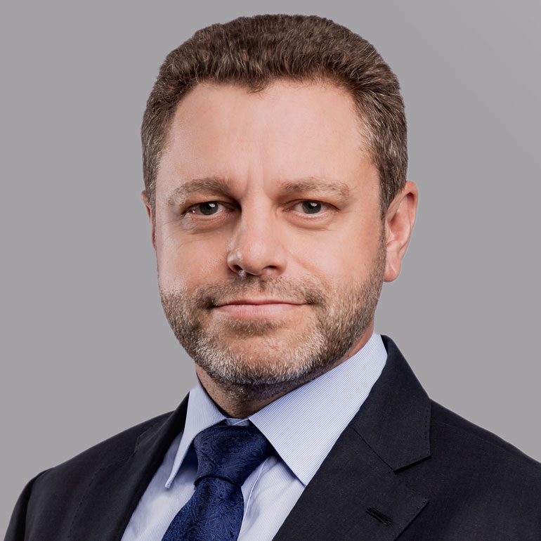 Porträt eines Mannes mit Kurzhaarschnitt und Bart, der einen eleganten Anzug und eine Krawatte trägt. Er hat einen neutralen Gesichtsausdruck und schaut direkt in die Kamera, vor einem grauen Hintergrund.