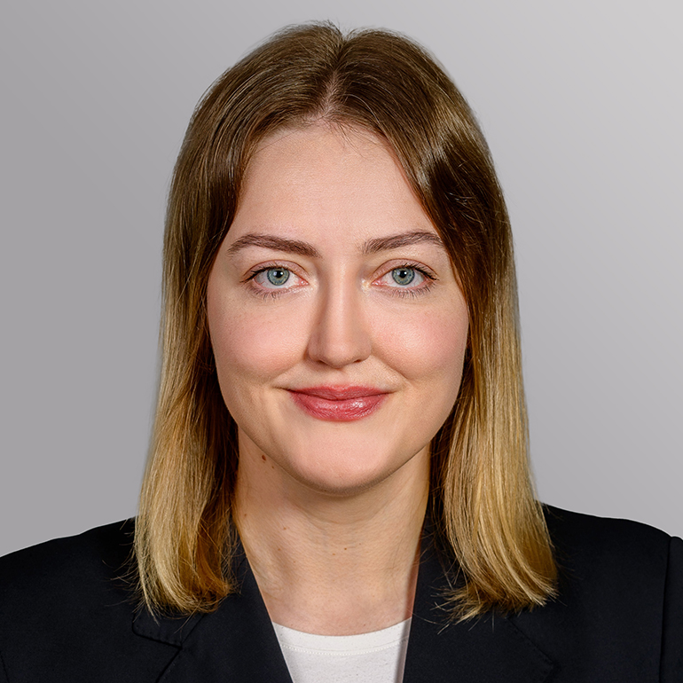 Ein Portrait einer jungen Frau mit blonden, schulterlangen Haaren und einem freundlichen Lächeln. Sie trägt einen schwarzen Blazer und hat einen neutralen Hintergrund. Ihre Augen sind blau und sie sieht direkt in die Kamera.
