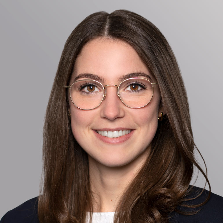 Ein Porträt einer Frau mit langen, braunen Haaren und einer freundlichen Ausstrahlung. Sie trägt eine Brille mit runder Fassung und hat ein Lächeln auf den Lippen. Der Hintergrund ist hellgrau. Sie ist in einem formalen Outfit gekleidet.